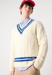 Maglione a trecce color crema con un bordo a V blu e banda inferiore a righe. Indossato su una camicia a righe blu chiaro.