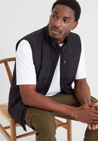 Black quilted vest with a high collar, closed with a zipper, worn over a white t-shirt. Olive green pants, sitting on a wooden chair.