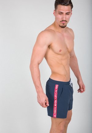 Navy swim shorts with elastic waistband and drawstring, featuring a red side stripe that says "REMOVE BEFORE FLIGHT."