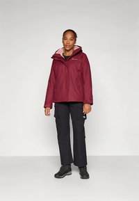 Bordeauxrode waterdichte jas met capuchon, ritssluiting aan de voorkant en roze voering, gecombineerd met zwarte cargobroek en zwarte wandelschoenen.