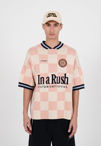 Model masculin purtând o cămașă cu mâneci scurte în carouri roz și crem, cu textul „In a Rush International” și o șapcă bej cu inscripția „Always Late Club.”