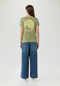 Olijfgroene katoenen t-shirt met een zonafbeelding en tekst op de rug, gecombineerd met wijde denim jeans en witte schoenen.