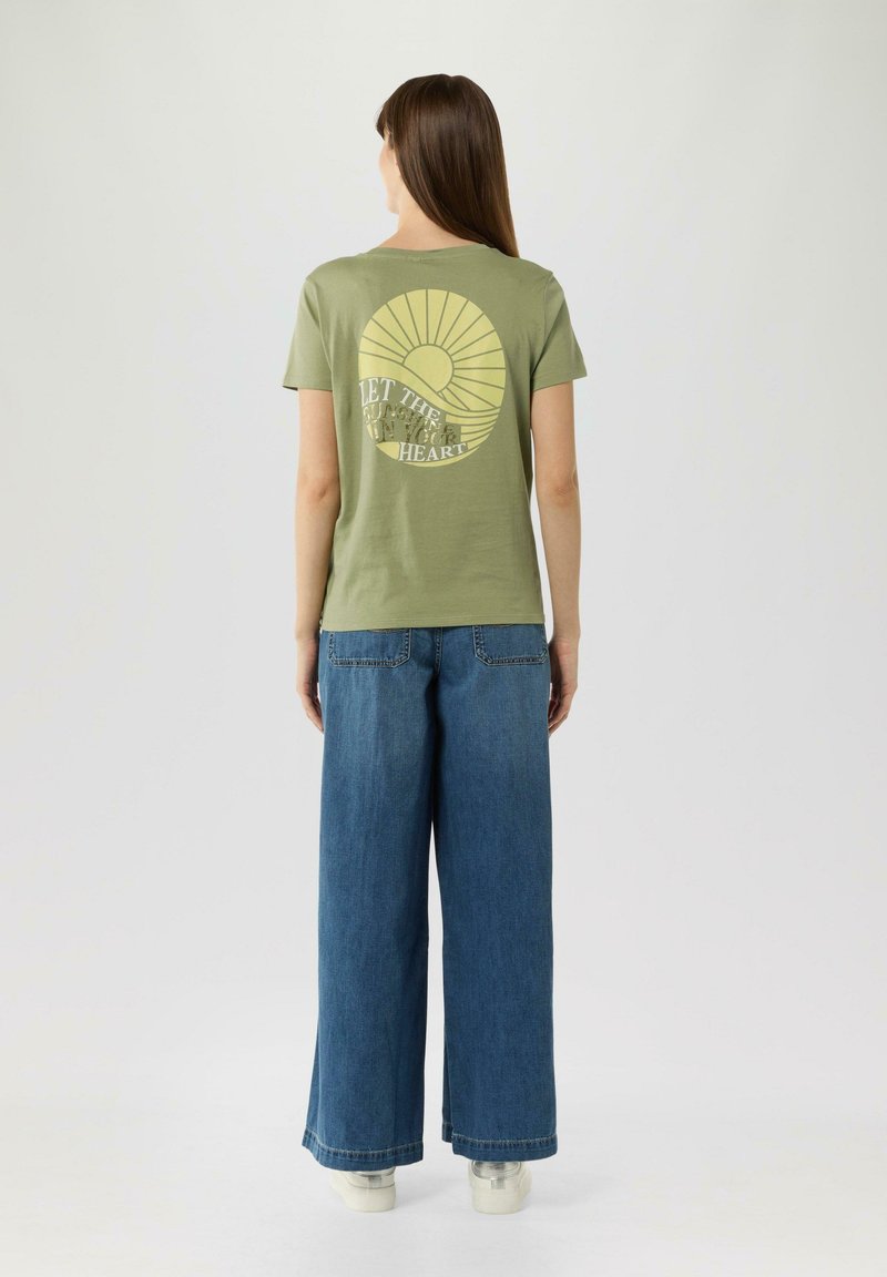 Olijfgroene katoenen t-shirt met een zonafbeelding en tekst op de rug, gecombineerd met wijde denim jeans en witte schoenen.