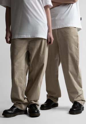 Kaks inimest, kes kannavad valgeid t-särke, beeže lahtiseid püksiseelikuid ja musti nahast paeltega kingi, seisavad heledal ühevärvilisel taustal.