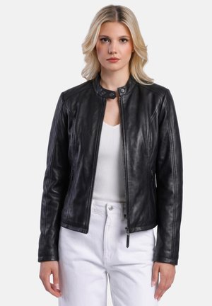 Mujer rubia con una chaqueta de cuero negra ajustada sobre un top blanco de canalé y pantalones blancos, posando frente a un fondo liso.