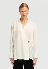 Weiße Wickelbluse mit langen Ärmeln, einem spitzen Kragen und silbernen Hardware-Details in der Taille. Glattes, leichtes Gewebe mit einem dezenten Glanz.