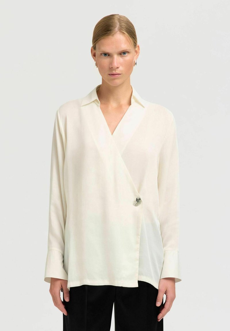 Weiße Wickelbluse mit langen Ärmeln, einem spitzen Kragen und silbernen Hardware-Details in der Taille. Glattes, leichtes Gewebe mit einem dezenten Glanz.