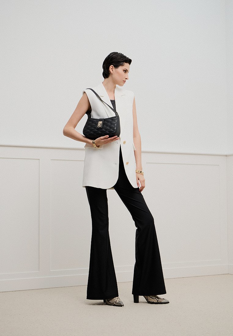 Borsa hobo nera con texture trapuntata, indossata con un blazer bianco senza maniche e pantaloni neri a zampa, su uno sfondo neutro.