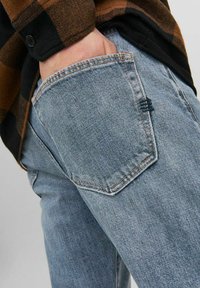 Jeans de denim azul claro con corte recto. Presentan un bolsillo trasero con un detalle de costura en zigzag y un ligero desvanecimiento. Se llevan con una camisa a cuadros.
