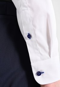 Manga de camisa blanca con botón azul y ojete a juego, con una textura suave y pliegues nítidos, situada sobre un fondo de tela oscura.