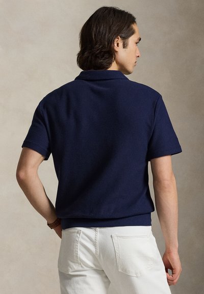 Άνδρας με μεσαίου μήκους σκούρα μαλλιά, φορώντας μπλε κοντομάνικη πικέ πόλο μπλούζα και λευκό παντελόνι, στέκεται μπροστά σε μπεζ φόντο.