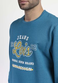 Sudadera en color teal con cuello redondo. Presenta texto impreso en blanco y amarillo: "JEANS Lois 1962 MARCA ORIGINAL DE DENIM ART&DESIGN."