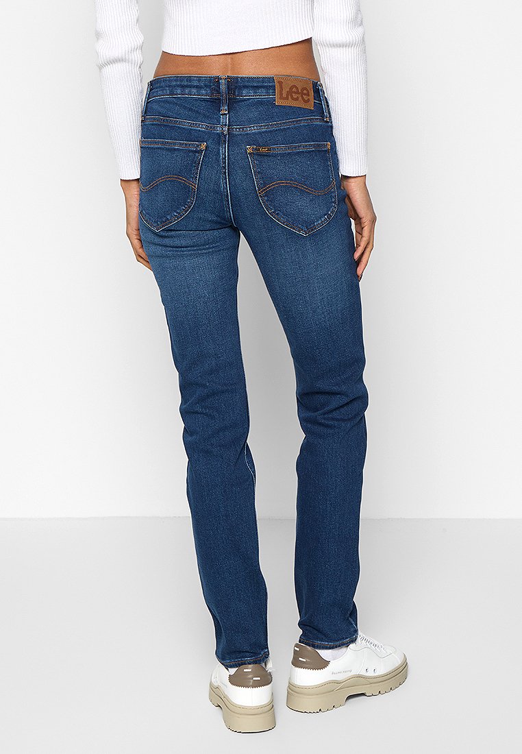 Femme portant un jean Lee bleu et des baskets blanches, vue de dos, se tenant contre un fond blanc uni.