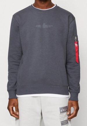 Mężczyzna ubrany w ciemnoszary sweter Alpha Industries z prążkowanymi mankietami i ściągaczem, z czerwoną metką na kieszeni rękawa oraz jasnoszare spodnie z łatami.