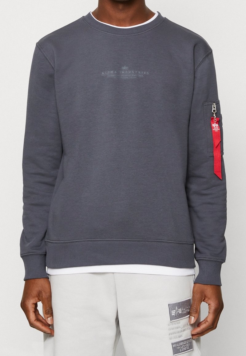 Homme portant un sweat-shirt gris foncé Alpha Industries avec des poignets et un bas côtelés, une étiquette rouge sur la poche de la manche, et un pantalon gris clair avec des patchs.