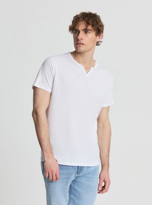 Mężczyzna z kręconymi włosami, ubrany w białą koszulkę henley z krótkim rękawem i jasnoniebieskie dżinsy, stoi na tle jednolitego jasnego tła.