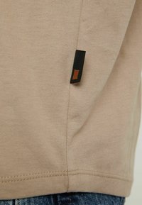 Détail d'un vêtement en tissu beige avec une petite étiquette rectangulaire noire et brune près de l'ourlet au-dessus d'un jean en denim.