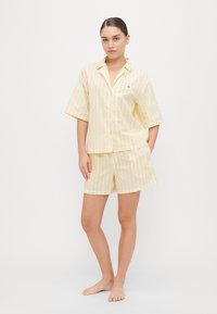 Ensemble de pyjama rayé jaune clair et blanc. Chemise à manches courtes avec boutons et shorts assortis. Tissu doux avec une coupe décontractée. Petit détail de logo.