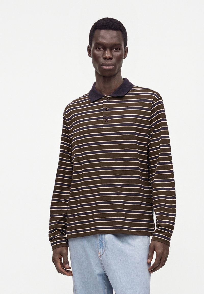 Jeune homme portant un polo marron rayé à manches longues et un jean bleu clair, debout devant un fond clair uni.