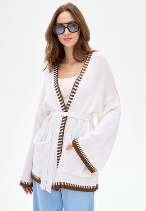 Kobieta w dużym białym dzianinowym kardiganie z brązową lamówką, białym paskiem, niebieskich spodniach i dużych ciemnych okularach przeciwsłonecznych, stojąca na tle białego tła.