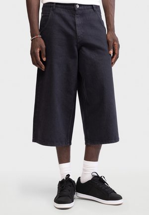 Bărbat purtând pantaloni scurți negri, largi, până la genunchi, șosete albe, pantofi sport negri și o brățară cu mărgele, stând în fața unui fundal simplu și deschis la culoare.