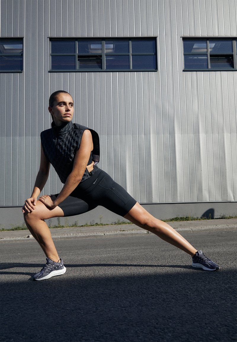 Chaleco sin mangas negro acolchado sobre unos pantalones cortos negros ajustados, acompañado de zapatillas de running oscuras con textura. El fondo presenta una pared gris corrugada.