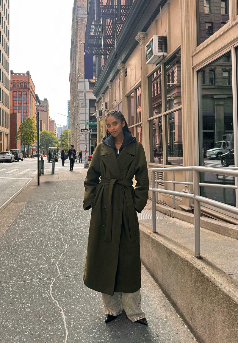 Dlhý olivovo zelený kabát s opaskom v páse, prehodený cez tmavú mikinu s kapucňou. V kombinácii s širokými svetlými džínami a čiernymi špicatými opätkami.