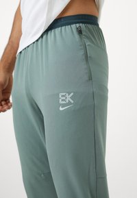 Sportovní kalhoty Nike ve světle zelené tkanině, s kapsou na zip, logovými detaily a elastickým pasem se značkou "NIKE."