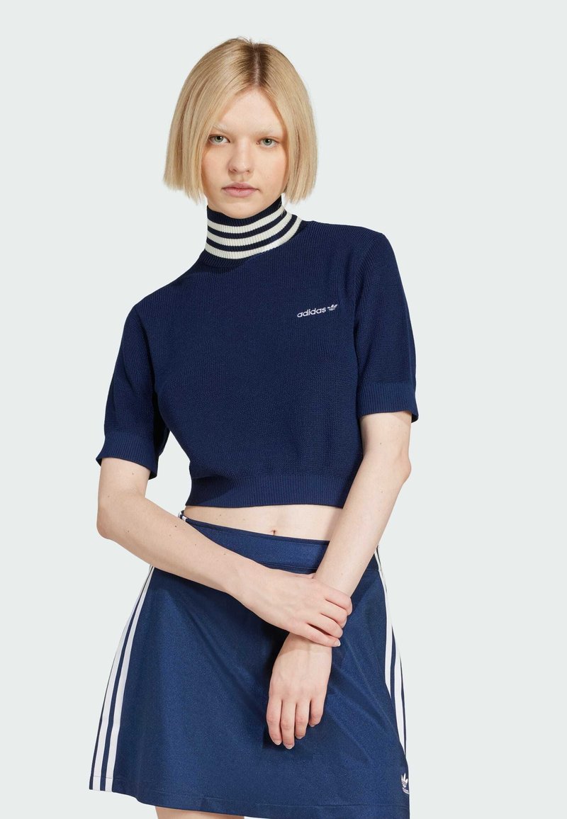 Pull cropped navy à manches courtes et col montant côtelé, avec des rayures blanches et logo Adidas brodé. Porté avec une jupe assortie.