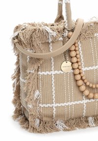 Beżowa torba typu tote wykonana z plecionego materiału, z białym wzorem, frędzlami na krawędziach oraz rączką z jasnobrązowej skóry ekologicznej. Ozdobiona dekoracyjnymi koralikami i posiada metkę.