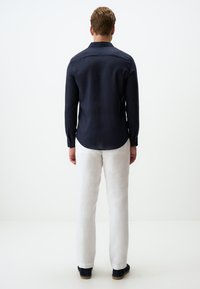 Navy lange mouw shirt met een button-down kraag, gecombineerd met witte linnen broek en donkere espadrilles, getoond vanuit de achterkant.