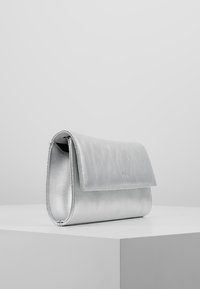 Silberne metallische rechteckige Handtasche mit Klappverschluss, auf einem weißen Podest vor einem schlichten hellgrauen Hintergrund ausgestellt.