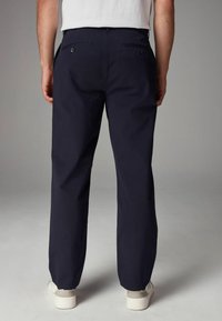 Un pantalon en coton bleu marine avec une coupe droite, des poches arrière avec des fermetures à bouton, associé à des chaussures à semelle blanche.