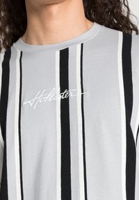 Grå randig t-shirt med vertikala svarta och vita ränder, rund halsringning och broderad "Hollister"-logotyp i vitt på bröstet.