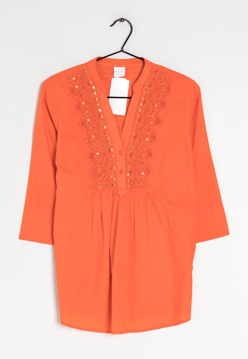 Camicia tunica arancione con scollatura a V, maniche a tre quarti, ricami decorativi e dettagli con bottoni sulla parte anteriore. Appesa a un gancio nero.