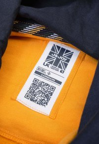 Tissu noir avec une doublure intérieure orange, comportant une étiquette blanche avec un code QR, le drapeau britannique, et du texte détaillant la marque et l'emplacement.