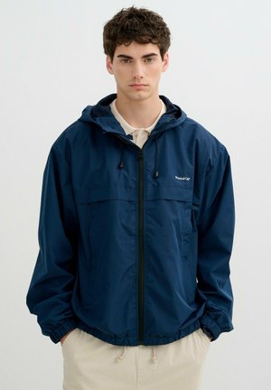 Joven con cabello rizado y oscuro, vestido con una cazadora cortavientos azul marino con capucha y pantalones de color claro sobre un fondo liso.