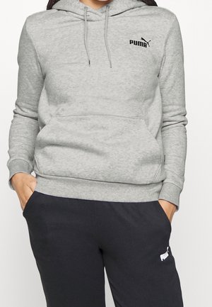 Kobieta w jasnoszarej bluzie z kapturem Puma z kieszenią na przodzie i czarnych spodniach dresowych Puma, ręce w kieszeniach, przycięty widok.