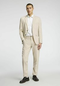 Beżowy garnitur o dopasowanym kroju, z pojedynczym zapięciem na guzik i klapami notch. Biała koszula pod spodem, w połączeniu z ciemnymi półbutami na sznurówki.