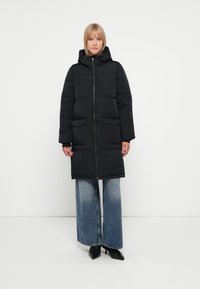 Manteau noir rembourré avec capuche, présentant une fermeture éclair à l'avant, deux grandes poches à rabat et une coupe longueur genou. Porté par-dessus un jean à jambes larges.