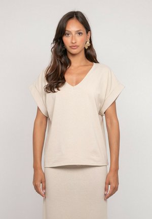 Femme aux longs cheveux foncés portant un haut beige à col en V et manches courtes ainsi qu'une jupe assortie, se tenant devant un fond clair uni.