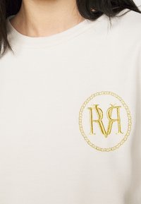 Vaaleanbeige puuvillapaita, jossa on pyöreä kultaisilla kirjontakirjaimilla varustettu logo, jossa on yhdessä kiedotut "R" ja "V" vasemmassa rinnassa.