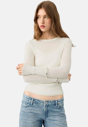 Dugi top s rebrastim uzorkom u nijansi nijanse bijele, s okruglim izrezom i manšetama s rupama za palce, uparen s visokim svijetloplavim trapericama.