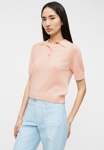 Πόλο μπλουζάκι με κοντά μανίκια σε απαλό παστέλ ροζ, με κλασικό γιακά, κούμπωμα με κουμπιά και ριμπ τελείωμα, σε συνδυασμό με ανοιχτόγαλα παντελόνι.