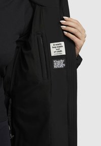 Käsi hoiab avatud musta jope, mille sisemistelt siltidelt on näha tekste "Kangast valmistatud soe kallistus" ja "Khujo originaal muudetavad niidid".