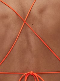 Neonové oranžové popruhy se kříží na hladkém, opáleném povrchu pleti, svázané v dolní části zad, čímž zdůrazňují minimalistický design bikiny.