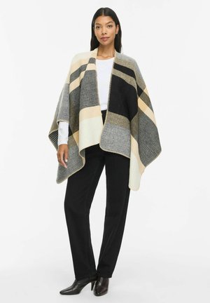 Femme portant un poncho rayé noir, beige et gris par-dessus un haut blanc avec un pantalon noir et des bottines noires à talons, debout sur fond blanc.
