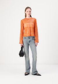Oranje pailletten top met lange mouwen en een doorschijnend ontwerp, gecombineerd met lichtblauwe gescheurde jeans en zwarte puntige ballerina's. Houdt een kleine zwarte handtas vast.