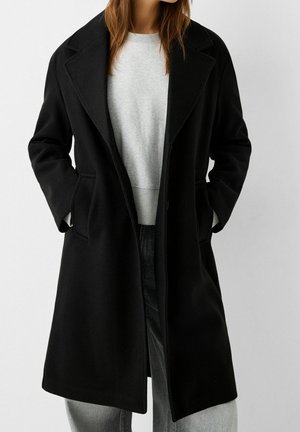 Femme portant un long manteau noir par-dessus un sweat-shirt gris clair et un pantalon gris, les mains dans les poches du manteau, sur un fond uni.