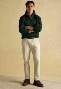 Młody mężczyzna stoi z założonymi rękami, ubrany w ciemnozielony sweter z krótkim zamkiem, beżowe spodnie, brązowy pasek i ciemno brązowe buty na tle jednolitym.